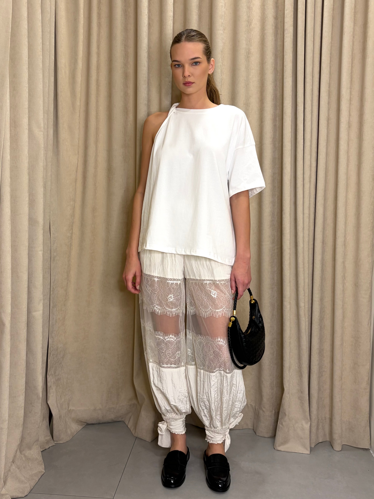 Woman wearing a white top and sheer pants standing against a beige curtain.